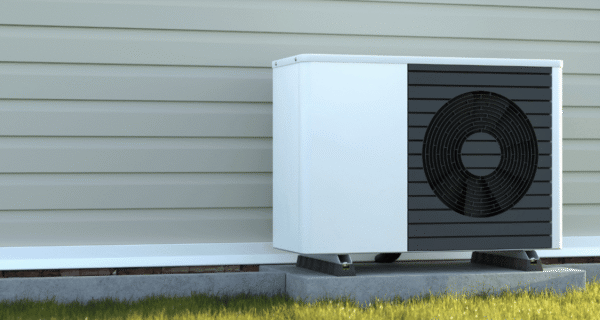 Outdoor air conditioner unit on a raised concrete pad beside beige siding, featuring a black grille and circular fan, with grass in the foreground.