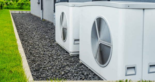 Two large white outdoor HVAC units with circular fans sit on a gravel strip along a gray-blue building, bordered by a green lawn on the left.