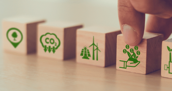 Row of wooden blocks with green eco icons: location pin with tree, CO2 cloud, wind turbines, a hand placing a plant with coins, and a factory.