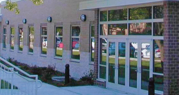 Exterior view of a light-colored building with rectangular windows, a glass entrance, brick column, and a white railing ramp along the walkway.