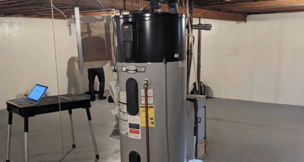 Basement utility room with a tall water heater, a work table with laptop, exposed beams, coiled hose on the right, and a person standing near heater.