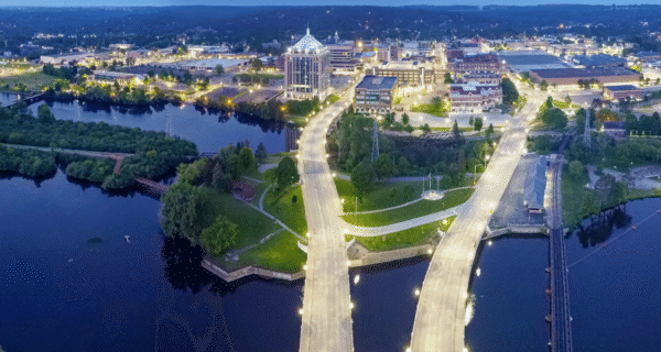 An aerial view of a riverfront city at dusk: two illuminated bridges span the water, with highways, green parks, and a cluster of lit buildings.
