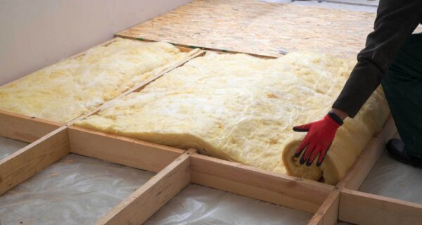 Person wearing red gloves places yellow fiberglass insulation between wooden floor joists at a construction site; OSB panels and framing visible.