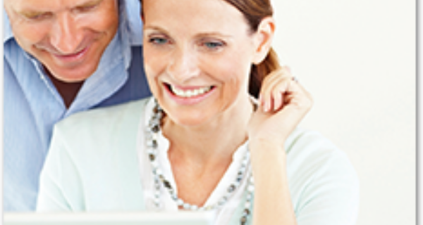 Two adults sit together and smile while looking at a laptop screen in a bright, white setting.