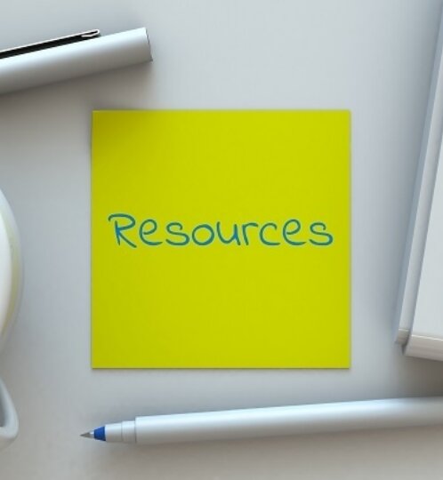 Yellow sticky note reading 'Resources' in blue, on a light desk with a coffee mug, a white keyboard, a white marker above, and a blue pen below.