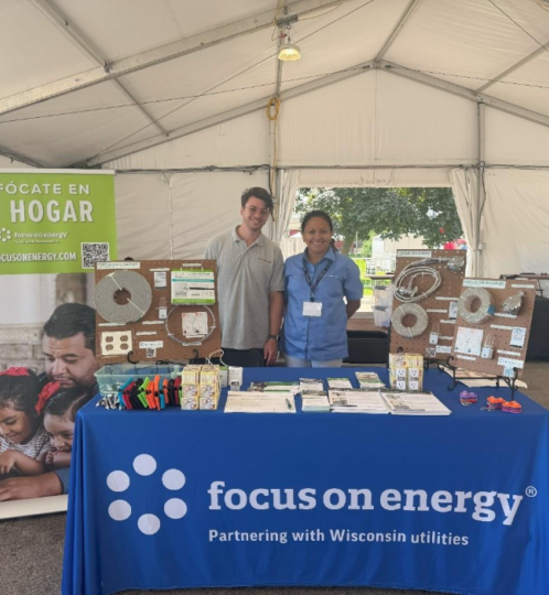 Two people stand behind a blue Focus on Energy table with display boards and brochures inside a large white event tent.