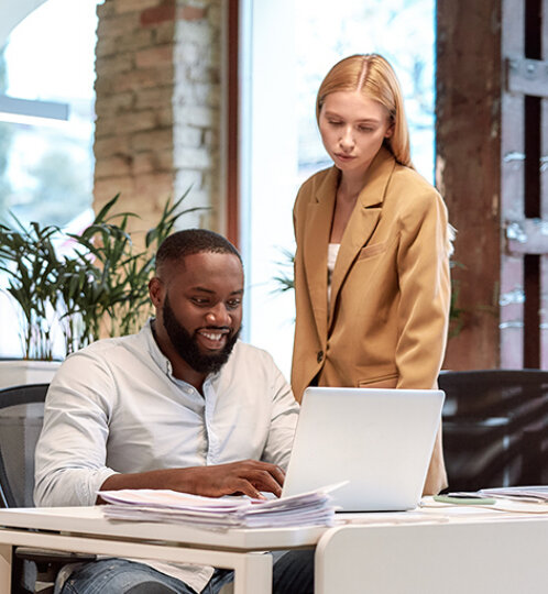 Two coworkers in a bright office: a person sits at a laptop with papers, while another stands beside, looking at the screen by brick walls and plants.