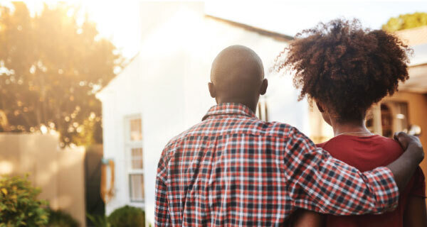 Two people walk arm-in-arm away from the camera toward a sunlit white house; one wears a red plaid shirt, the other has a curly afro.