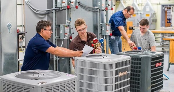 Four technicians in a workshop inspect an HVAC system around outdoor condenser units, holding papers and adjusting gauges.