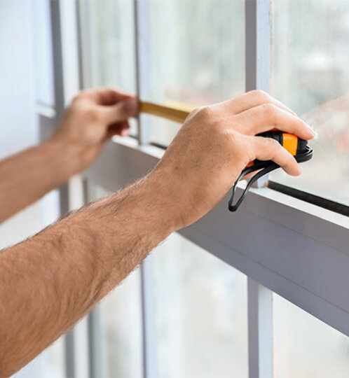 Close-up of hands measuring a window frame with a tape measure and a wooden shim, indoors by a glass window.