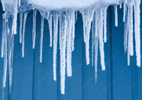 Snow-covered edge with numerous icicles hanging down, frost coating their tips, against a blue corrugated metal wall.