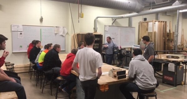 A group sits around a large woodworking workbench in a shop, listening to a presenter near a whiteboard with notes, with wood and tools in view.