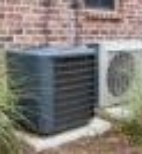 Two outdoor air conditioning condenser units on concrete pads beside a brick wall, with soil and greenery nearby.